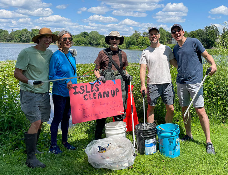 Five adults pose at a lakeshore with litter-picking tools, collection buckets, a bag of debris, and an &ldquo;ISLES CLEANUP&rdquo; sign