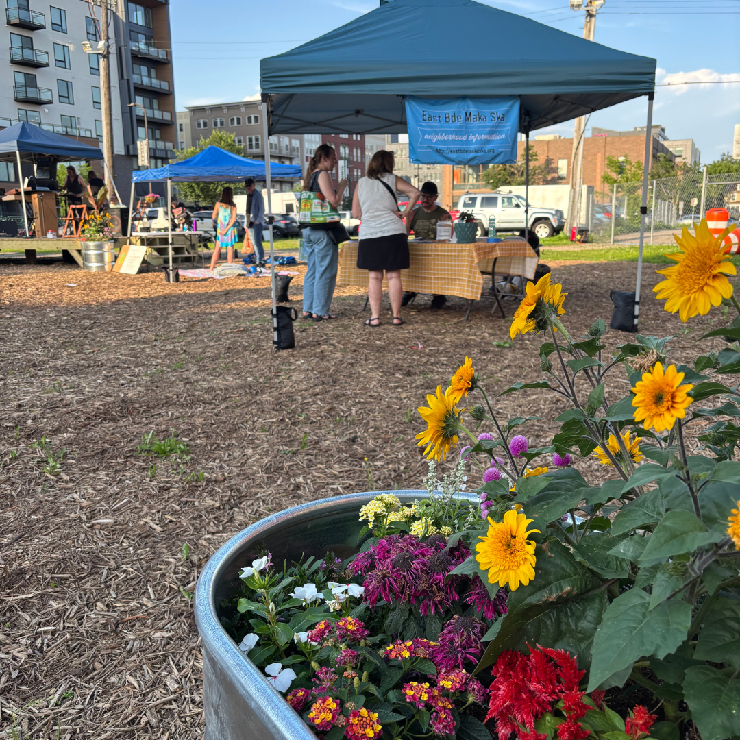 East Bde Maka Ska neighborhood organization table at Uptown Farmer Market a large foreground planter filled with colorful flowers