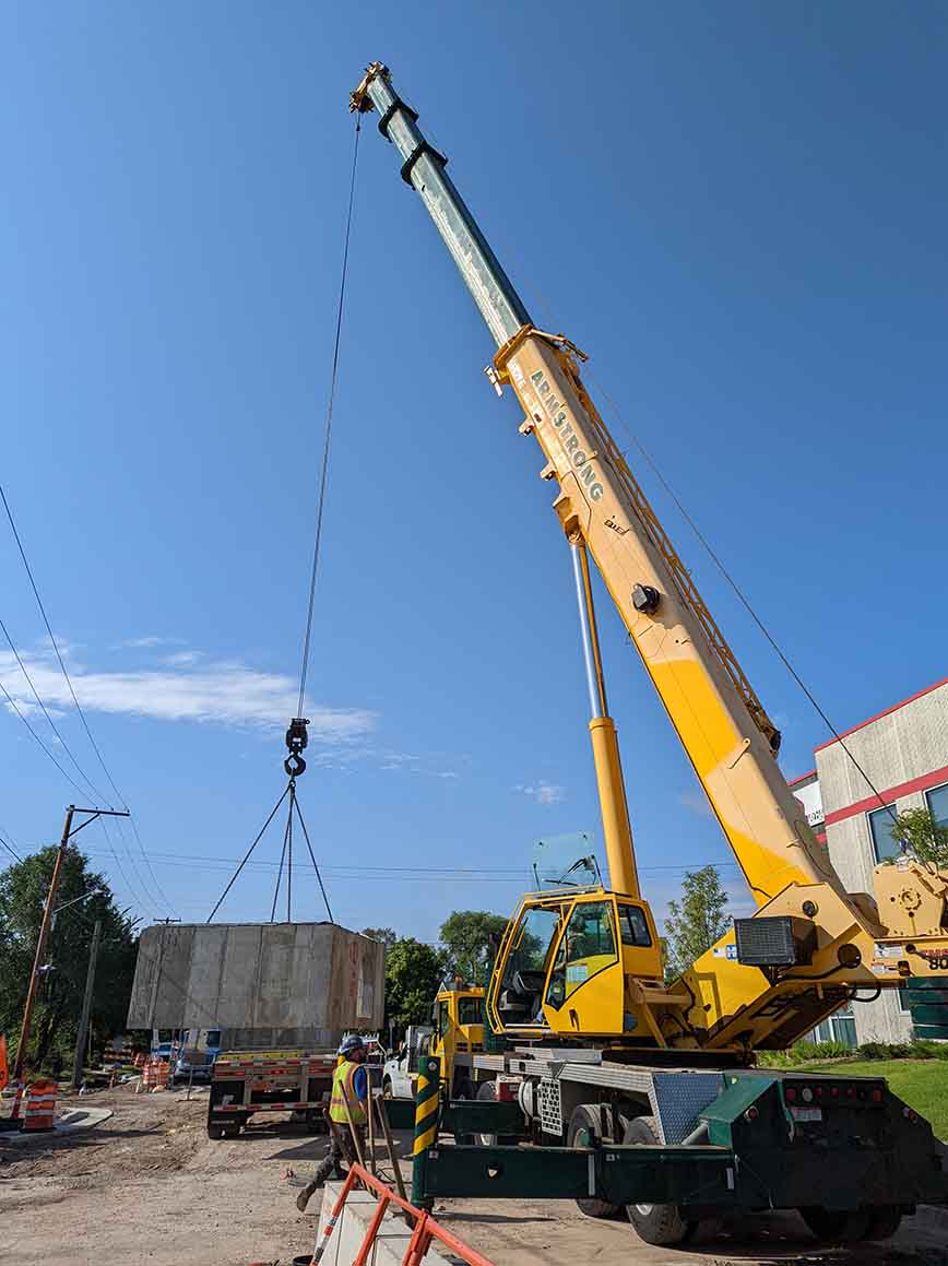 A crane lifts a concrete box structure off of a truck and prepares for installation. 