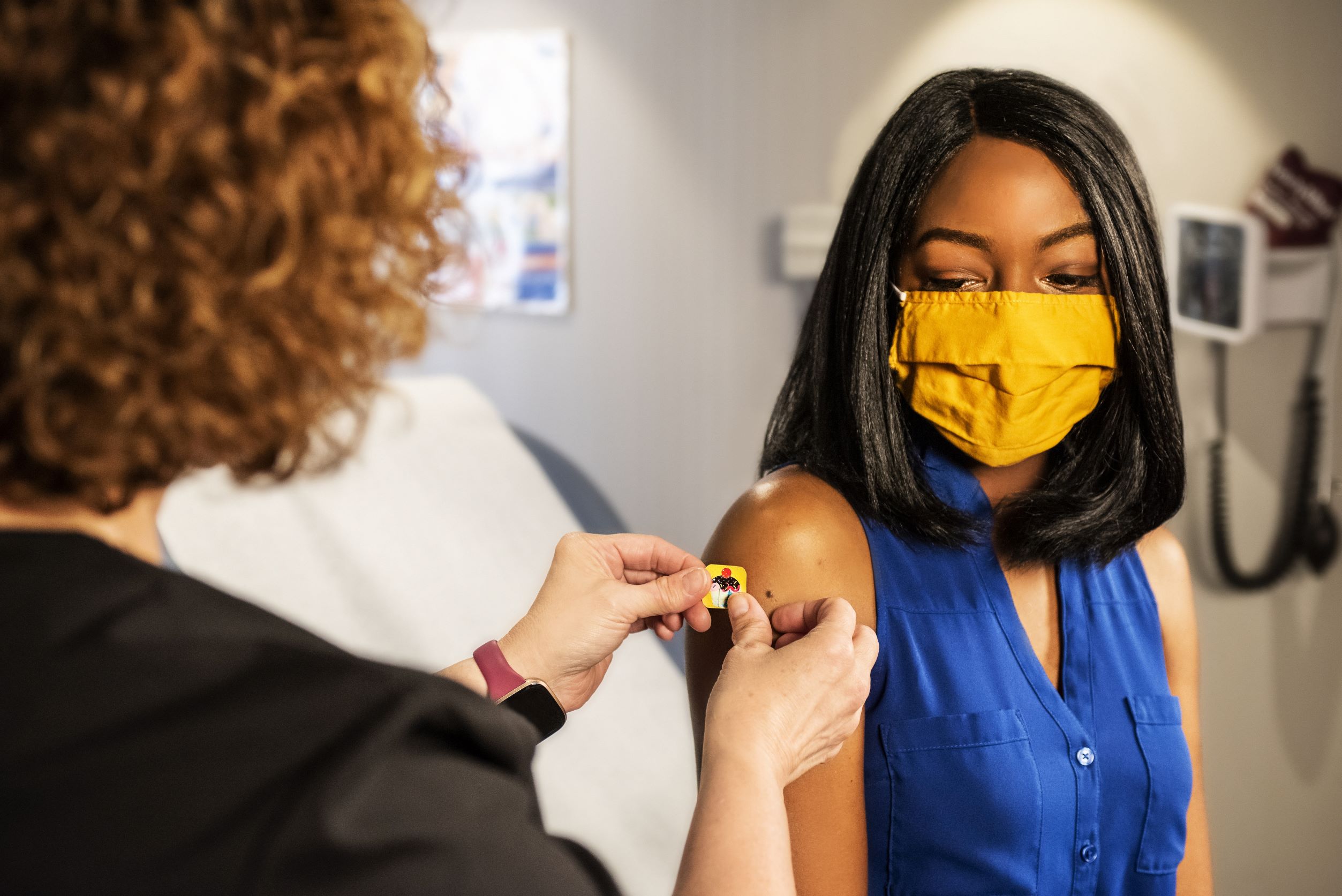 Young African American student with yellow mask receives a vaccine or shot in arm by a medical professional nurse