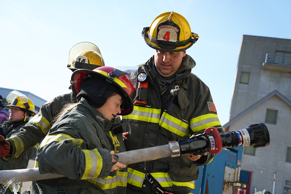 A community member participates in a hose demonstration with a Minneapolis firefighter
